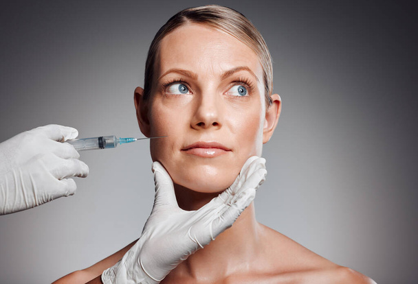 Beautiful mature woman getting botox injection in studio against a grey background. - Photo, image