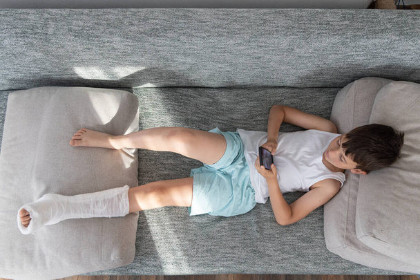 Μικρό αγόρι με σπασμένο πόδι σε γύψο, ξαπλωμένο στον καναπέ στο σπίτι, παίζοντας στο κινητό τηλέφωνο - Φωτογραφία, εικόνα