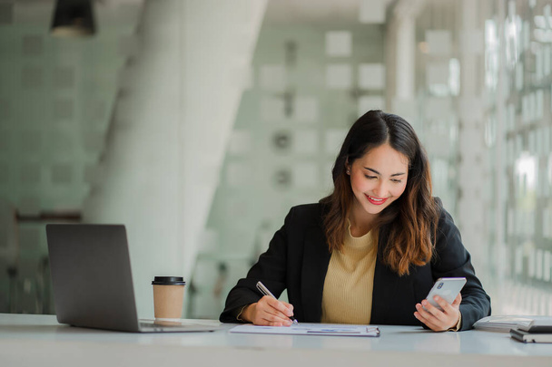 Úspěšná mladá asijská podnikatelka držící smartphone pracující na notebooku finanční graf data na stole v kanceláři. - Fotografie, Obrázek