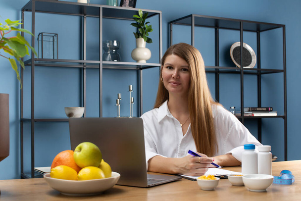 A female nutritionist smiles at the camera, sitting at her desk in a white shirt at a laptop, fresh fruits and food additives on the table. - Zdjęcie, obraz