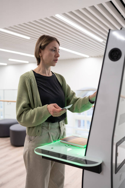 Middle-aged scandinavian woman using library self-service machine for borrow or return book, female holding ID card and clicking screen with finger, interacting with new smart library technologies - Foto, immagini