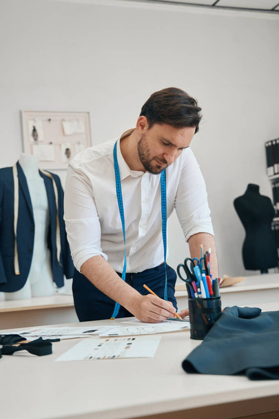 Klidný módní návrhář stojí u pracovního stolu s tužkou v ruce, zatímco pohled je zaměřen na list papíru se vzorem - Fotografie, Obrázek