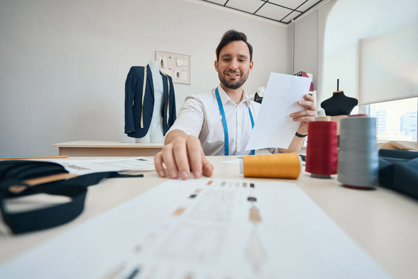 Радостный модельер сидит в окружении швейного оборудования, держа лист бумаги с рисунком будущей одежды в руке - Фото, изображение