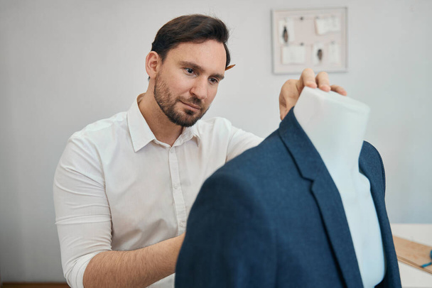 Focused fashion designer stands near mannequin while with his right hand makes professional work with product - Zdjęcie, obraz