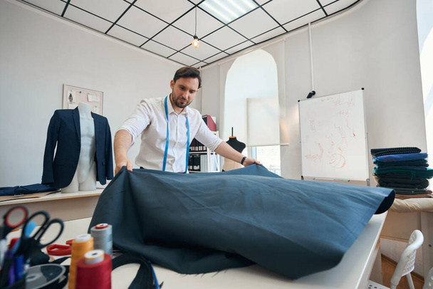 Diseñador de moda concentrado se para cerca de escritorio en taller de costura mientras se despliega rollo de tela en el escritorio - Foto, Imagen
