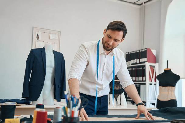 Kalme mode-ontwerper maakt voorbereidingen voor het maken van kleding terwijl naaien apparatuur ligt op de tafel voor hem - Foto, afbeelding