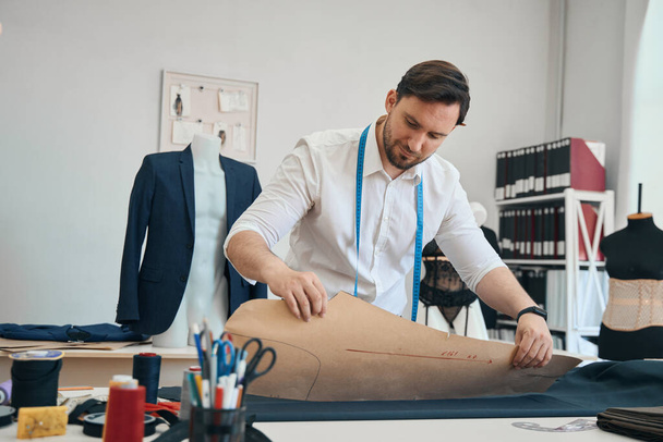 Calm fashion designer is applying blank of future clothes to fabric while behind him stands a mannequin with a jacket - Photo, image