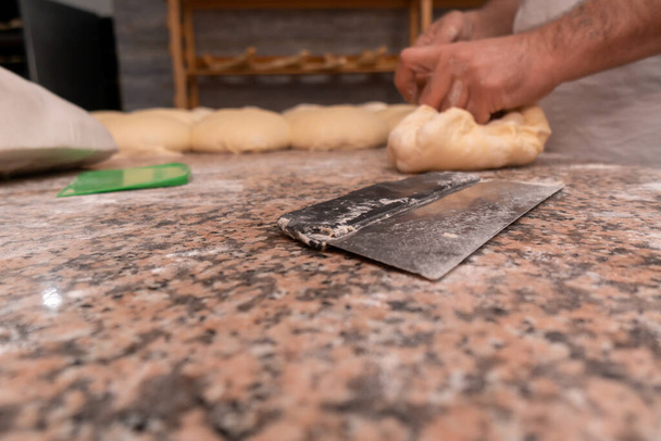 Close-up of a knife cutter for dough, in the background men's hands are kneading dough, there is a lot of prepared dough on the table for baking buns or other bakery products - Photo, image