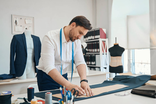 Вдохновленный молодой дизайнер стоит в швейной комнате, а на столе перед ним готовая ткань для работы - Фото, изображение