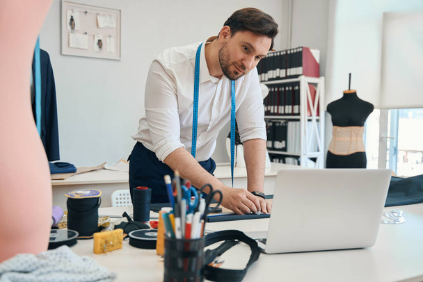 Συγκεντρωμένος σχεδιαστής μόδας κοιτάζοντας την οθόνη gadget στην επιφάνεια εργασίας, ενώ καθοδηγεί τη σιλουέτα του προϊόντος στο ύφασμα με τα χέρια στο εργαστήριο ραπτικής - Φωτογραφία, εικόνα