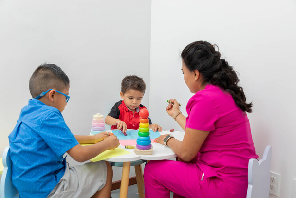 Latin çocuk doktoru, ofisinde oyuncaklarla masada oynayan iki çocukla oynuyor.. - Fotoğraf, Görsel