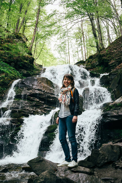 Happy smiling tourist girl with backpack near to cold fresh waterfall. Waterfall Shypit, cascade in Pylypets in forest. Carpathian Mountains, Ukraine. - Фото, зображення