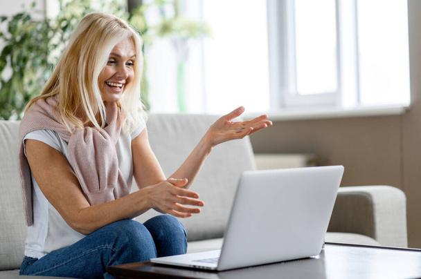 Happy friendly middle aged caucasian blonde woman in casual clothes, sitting on cozy sofa in living room, talking on video conference with family, friends or colleague, using laptop, smiling - Zdjęcie, obraz