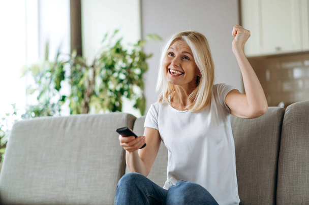 Leisure and entertainment concept. Positive excited caucasian blonde woman watching TV show or film, holding remote control, changes channels, while sitting on cozy sofa at home in living room, smiles - Foto, Bild