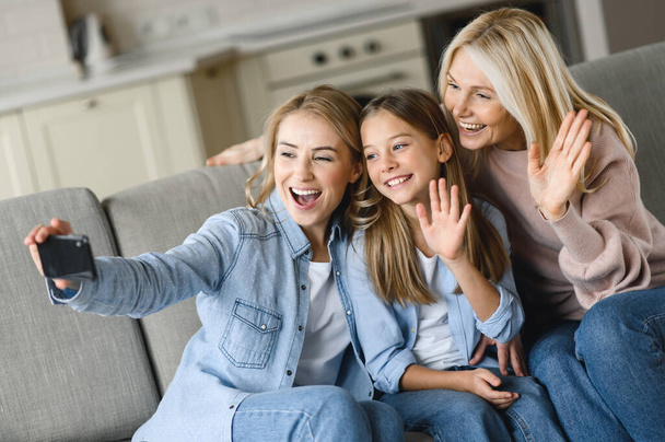 Three generations of Caucasian females. Happy grandmother, daughter and granddaughter, sitting on sofa in living room, having fun together, taking selfie posing for the camera, talking on a video call - Photo, image
