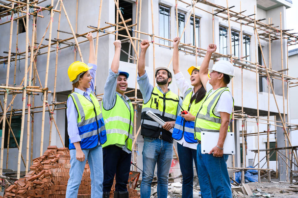 Skupina dodavatelů, inženýrů a formátů v bezpečnostních vestách s přilbami ruce nahoru, aby společně úspěšně dokončit stavební projekt na staveništi. koncepce spolupráce a úspěchu - Fotografie, Obrázek
