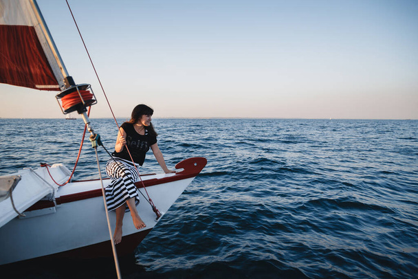 Young pretty woman in black shirt and striped skirt at luxury yacht in sea, sunset. - Foto, imagen