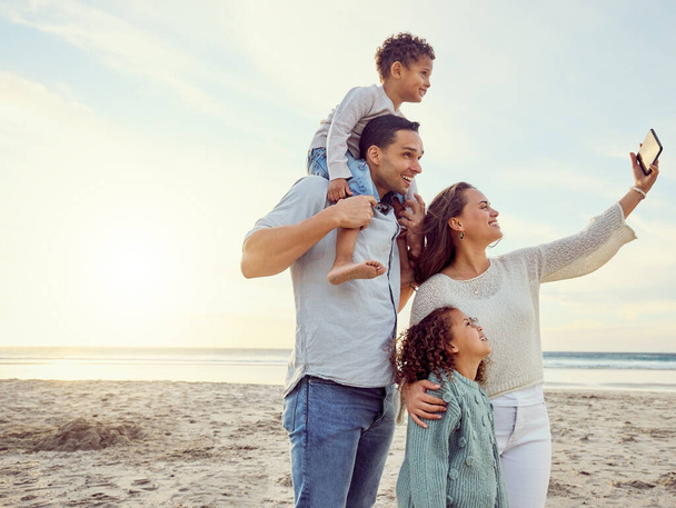 Jeune heureux mixte famille souriant pour un selfie debout à la plage ensemble. Femme hispanique souriante prenant une photo avec ses enfants et son mari avec son téléphone portable collant en vacances. - Photo, image