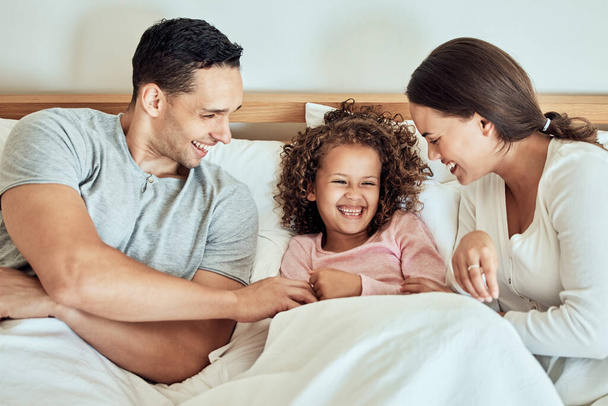 Playful parents tickling their little daughter while lying in bed together. Cheerful couple bonding with their adorable little girl at home in the morning. Cute little girl laughing while playing with - Foto, Imagen