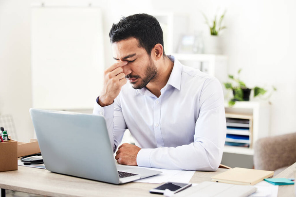 Young mixed race businessman suffering from a headache working on a laptop in an office alone at work. Hispanic man looking worried and upset while working. Male boss feeling tired sitting at a desk. - Photo, Image