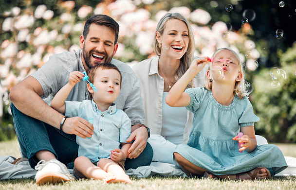 Lächelnde kaukasische Familie pustet Seifenblasen zum Spaß, während sie es sich an einem sonnigen Tag im Park oder Garten draußen gemütlich macht. Liebevolle Eltern, die mit verspielten Kindern eine unbeschwerte, glückliche Kindheit genießen - Foto, Bild