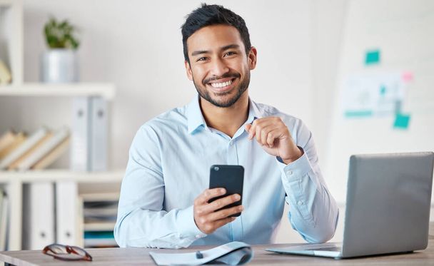 Πορτρέτο ενός νεαρού όμορφου χαρούμενου επιχειρηματία μικτής φυλής που χρησιμοποιεί τα μέσα κοινωνικής δικτύωσης στο τηλέφωνό του ενώ εργάζεται μόνος του σε ένα φορητό υπολογιστή σε ένα γραφείο στη δουλειά. Ένας ισπανόφωνος άντρας αφεντικό χαμογελάει χρησιμοποιώντας ένα κινητό στη δουλειά. - Φωτογραφία, εικόνα