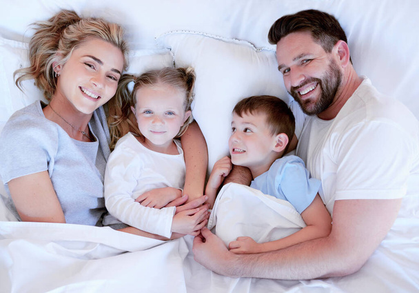 Above portrait view of a young family lying in bed. Married couple enjoying free time with their son and daughter at home. Adorable little girl and boy bonding with mother and father in the morning. - 写真・画像