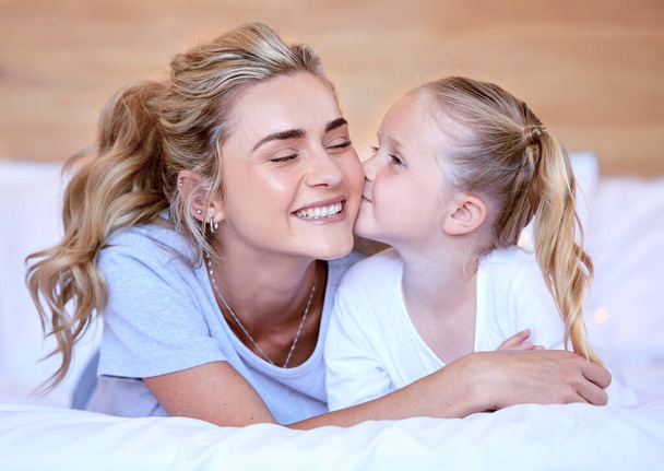 Two blonde caucasian females only looking relaxed and positive while lying in bed together. Young single mother kissing her adorable little daughter on an comfortable bed in a bright bedroom. - Foto, afbeelding