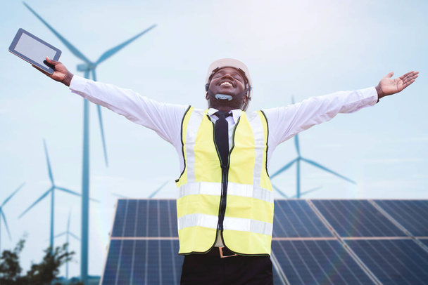 Επιτυχημένος αφρικανός μηχανικός διευθυντής σταθεί κρατώντας tablet μπροστά ηλιακή φωτοβολταϊκά και ανεμογεννήτριες παραγωγής ηλεκτρικής ενέργειας σταθμό. Έννοια της ανάπτυξης αειφορίας μέσω εναλλακτικής ενέργειας - Φωτογραφία, εικόνα