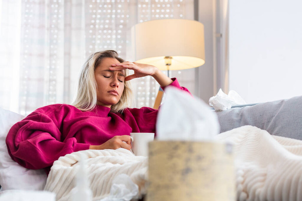 Sick woman with headache sitting under the blanket. Sick woman with seasonal infections, flu, allergy lying in bed. Sick woman covered with a blanket lying in bed with high fever, Coronavirus symptoms - Foto, Imagen