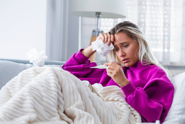 Sickness, seasonal virus problem concept. Woman being sick having flu lying on sofa looking at temperature on thermometer. Sick woman lying in bed with high fever. Cold flu and migraine. - Foto, Bild