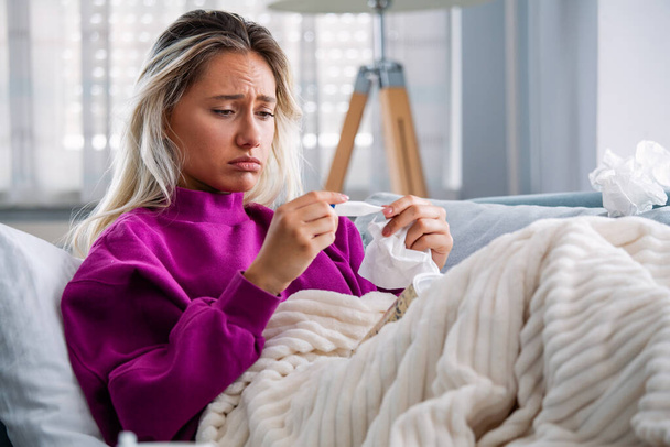 Sickness, seasonal virus problem concept. Woman being sick having flu lying on sofa looking at temperature on thermometer. Sick woman lying in bed with high fever. Cold flu and migraine. - Photo, Image