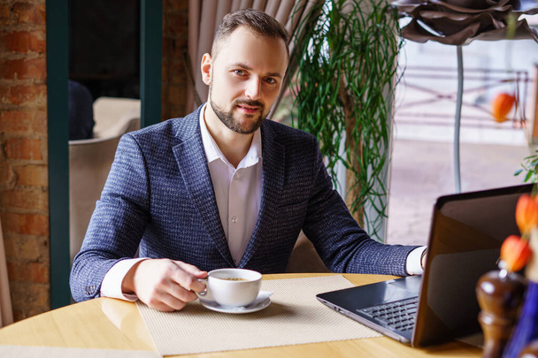 A young man in a business suit sits at a table in a restaurant. Businessman drinking coffee while working at his laptop in a restaurant or cafe - Foto, imagen