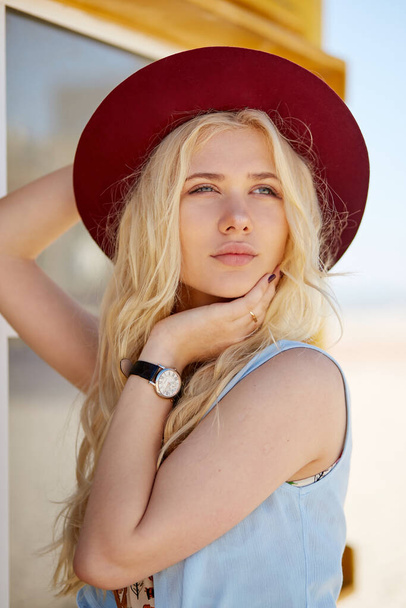 Profilkép egy szőke, elegáns lány göndör haj öltözött könnyű ruha és kalap, nézi a kamera, tengerparti háttér. - Fotó, kép