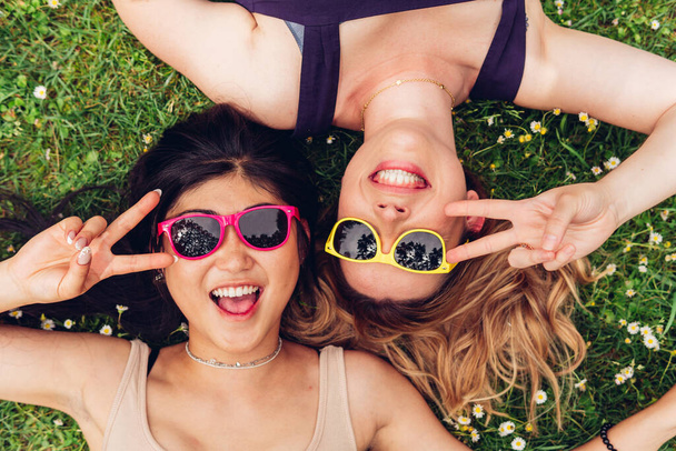 Top view of diverse women in trendy sunglasses gesturing V sign and looking at camera with smile while lying on blooming grass in park together - Fotoğraf, Görsel