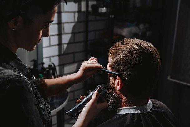 Mujer joven peluquero haciendo corte de pelo de hombre barbudo en la barbería. Autocuidado, belleza masculina. - Foto, Imagen
