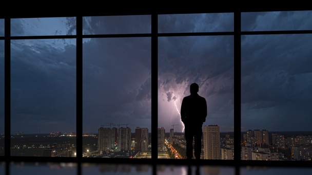 Чоловік стоїть біля панорамного вікна на фоні нічної блискавки
 - Фото, зображення