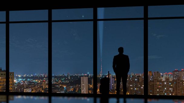 The man with a suitcase standing near a panoramic window against an evening city - Foto, imagen