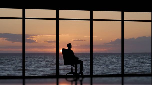 De man die bij het panoramische raam zit tegen de zonsondergang aan - Foto, afbeelding
