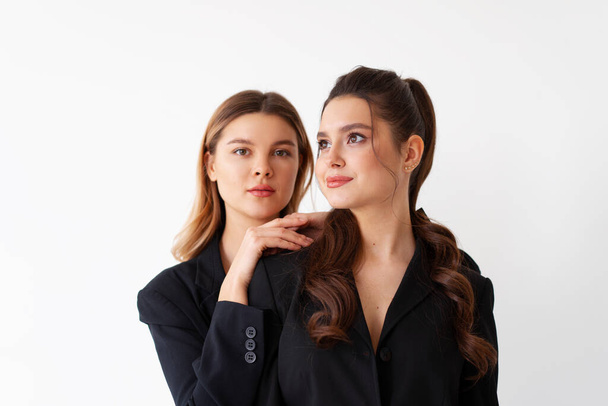 Concept of partnership in business. Two Young businesswoman dressed black formal suit standing studio isolated white background. Blonde woman put her hands on the shoulders of her friend. - 写真・画像