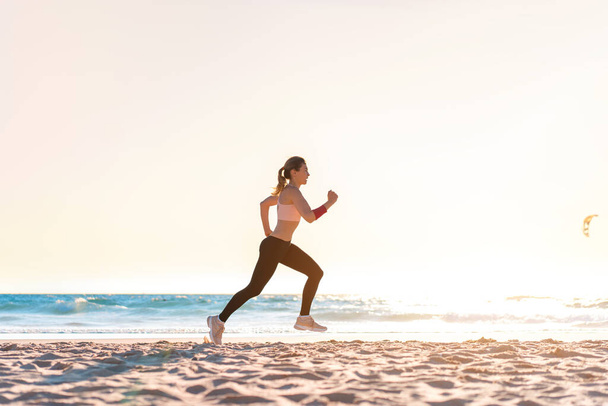 Sporty blonde woman running ocean beach. Young caucasian female exercising outdoors running seashore. Concept of healthy running and outdoors exercise. Active, sporty athlete jogging. Summer active - Photo, Image