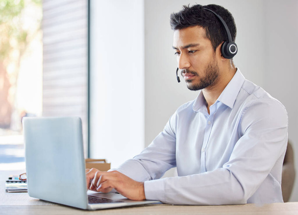 Businessman working in a call center. Operator working on his laptop. Young multiracial man working, wearing a headset. Young businessman working in customer service, on a call - 写真・画像