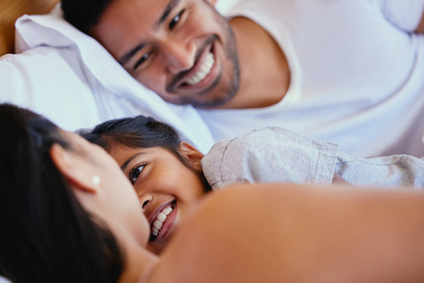 Adorable little girl smiling while lying in bed with her parents. Mixed race family with one child relaxing and bonding in the morning while still lying in bedroom. - Photo, image