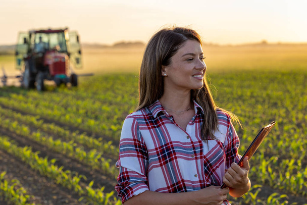 Portrait d'une jolie jeune agricultrice tenant un bloc-notes devant un tracteur dans un champ de maïs au printemps - Photo, image