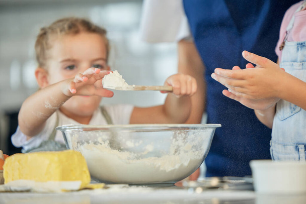 Little girl holding a spoon of flour. Family baking together at home. Two sisters baking with a parent. Caucasian girl putting flour into a bowl. Mother baking with her daughters. Siblings baking - 写真・画像