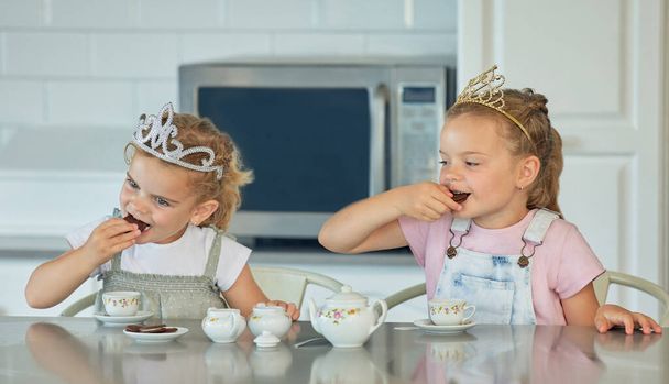 Two little girls having a princess tea party at home. Sibling sister friends wearing tiaras while playing with tea set and having cookies at kitchen table. Sisters getting along and playing together. - Foto, Bild