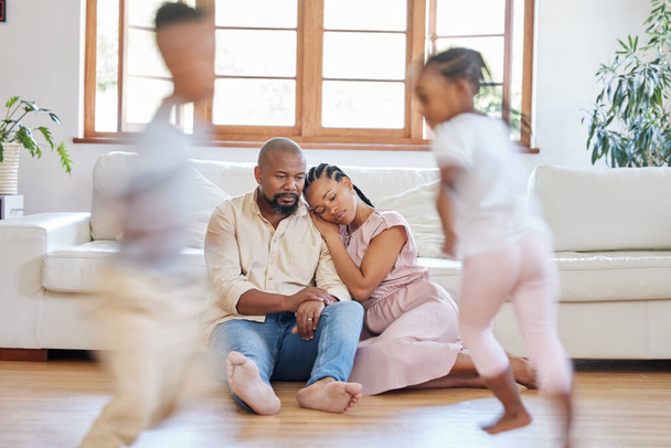 Tired African American couple sitting on the lounge floor looking tired and drained while their energetic children plays around them. Two black siblings having fun while their exhausted parents relax. - Фото, изображение