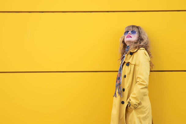 middle aged woman with yellow trench coat on yellow background in the street with copy-space - Foto, imagen