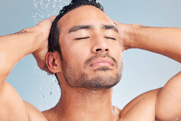 Close up of muscular asian man showering alone and washing his hair against a blue studio background. Fit and strong mixed race man standing under pouring water. Hispanic athlete enjoying hot shower - 写真・画像