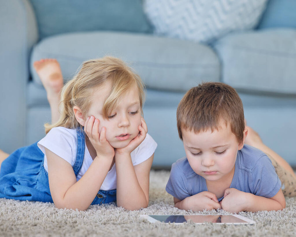 Young caucasian boy and girl watching cartoon videos online and playing fun games on digital tablet to learn from educational apps while lying on the carpet floor at home. Two kids using smart gadget. - Foto, Imagem